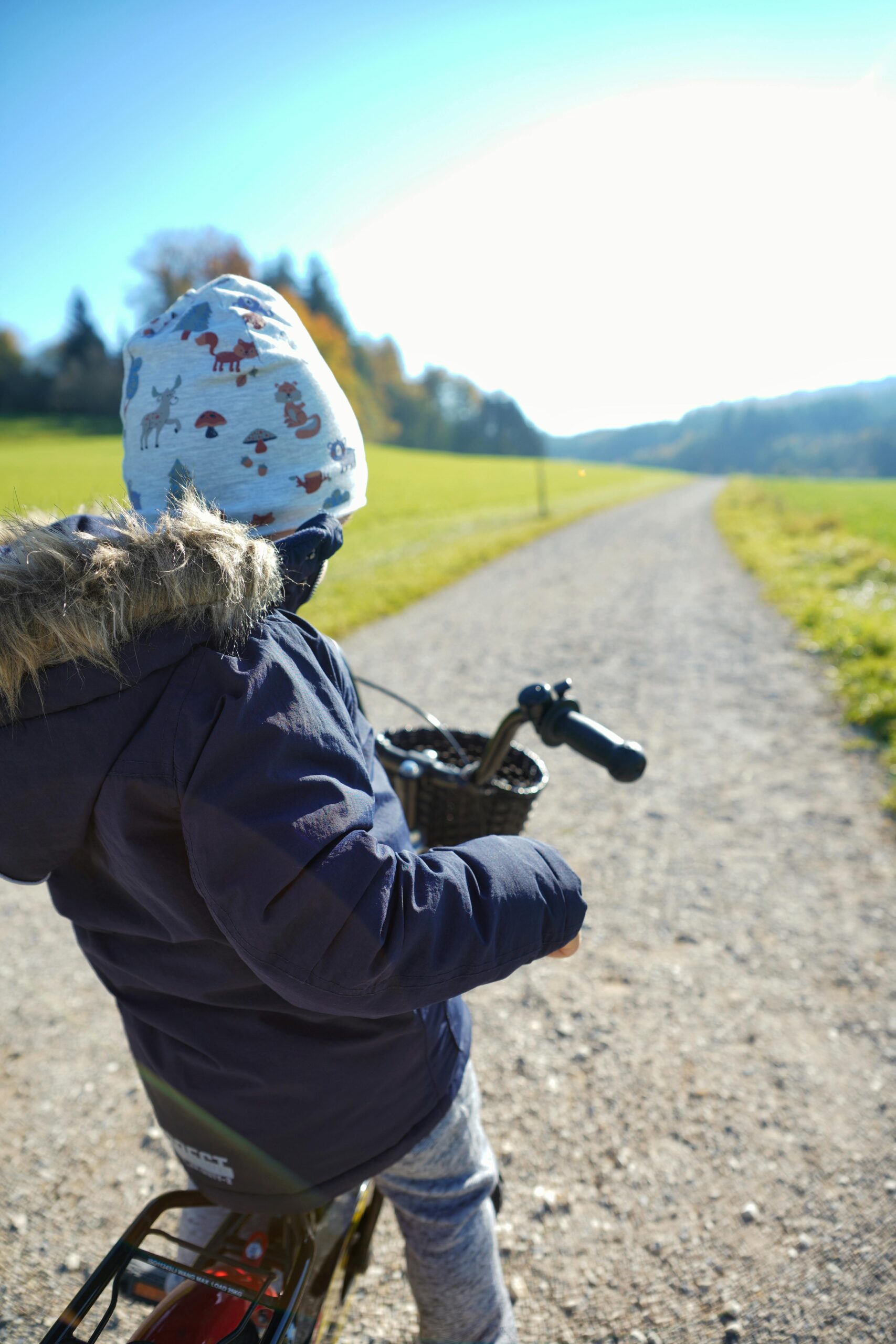Home A child in a jacket rides a bicycle on a sunny dirt path, enjoying the outdoors.